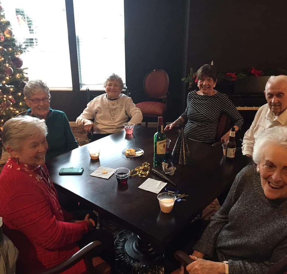 A group of elderly people enjoying a gathering indoors around a table with drinks and snacks.
