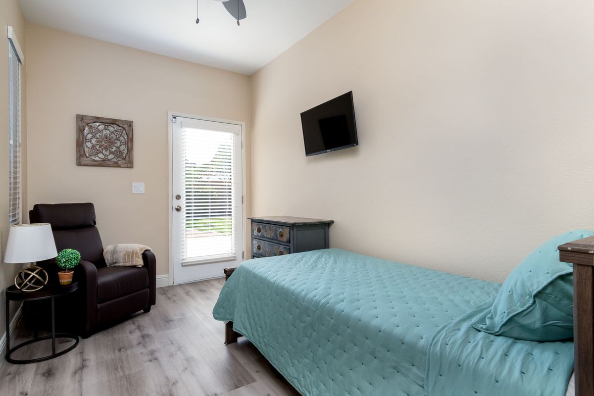 A cozy bedroom featuring a single bed with blue bedding, a wall-mounted TV, and an armchair next to a windowed door.