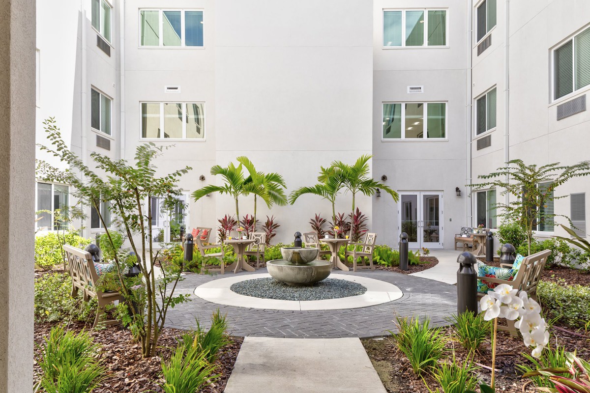 A modern courtyard with a central fountain, surrounded by plants and palms, enclosed by a building.