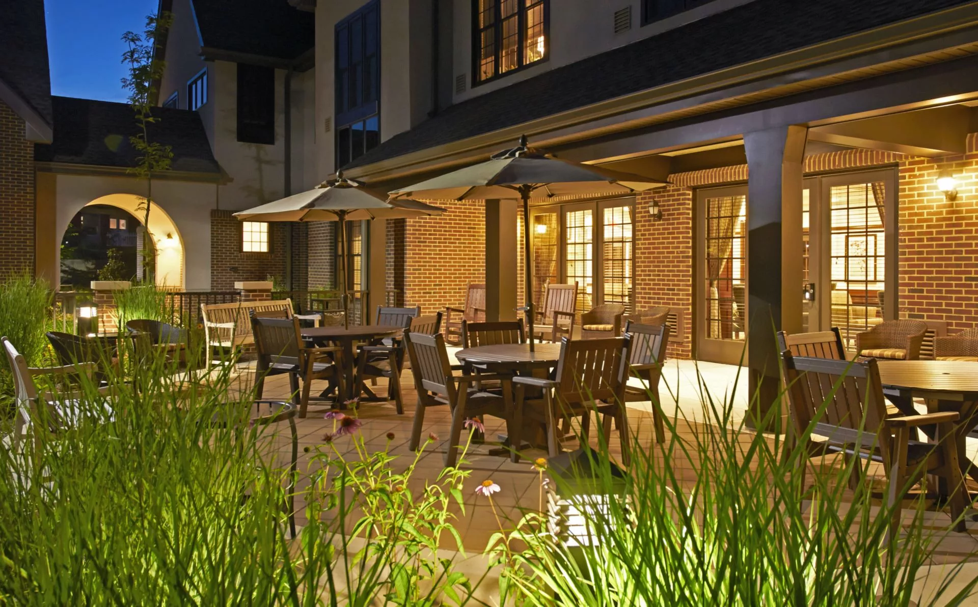 A cozy patio with tables and chairs under umbrellas, illuminated in the evening light.