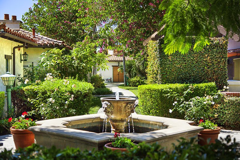 A tranquil garden scene featuring a central fountain surrounded by lush greenery and a pathway leading to a charming building.