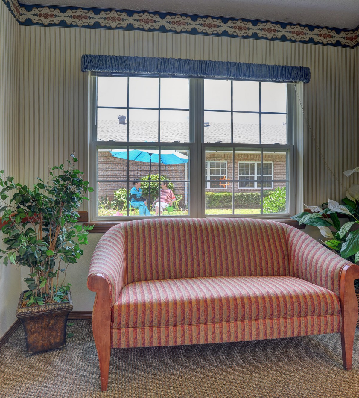 A cozy living room with a striped sofa, framed by large windows and indoor plants.