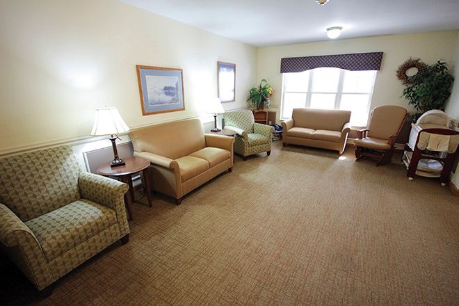A cozy living room with multiple sofas and chairs arranged around a central carpeted area, complemented by lamps and windows.