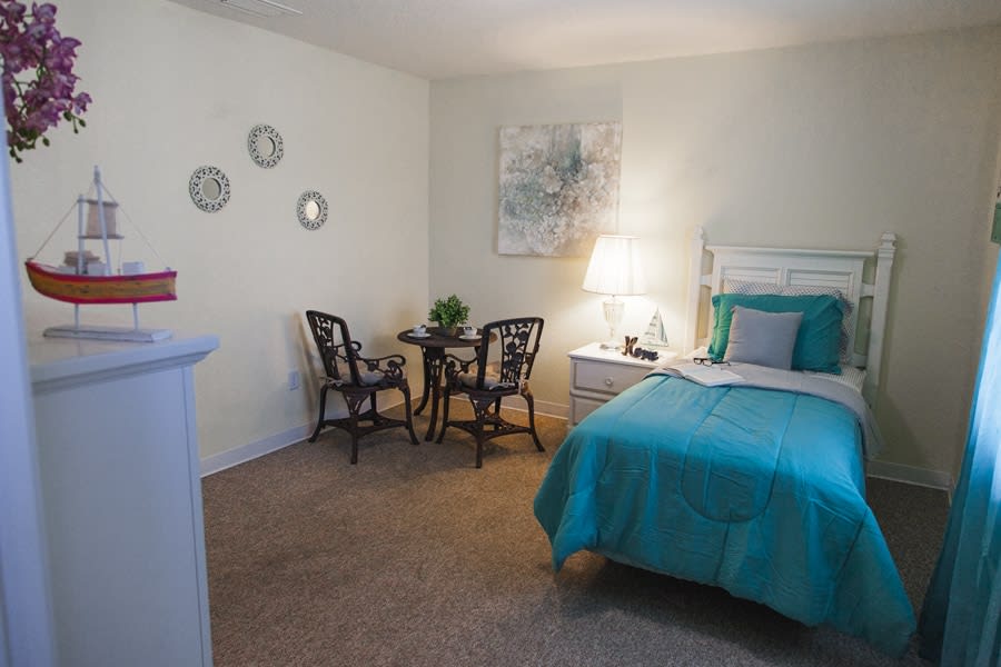 A cozy bedroom with a turquoise bedspread, a small table and two chairs, a lamp, and wall decor.