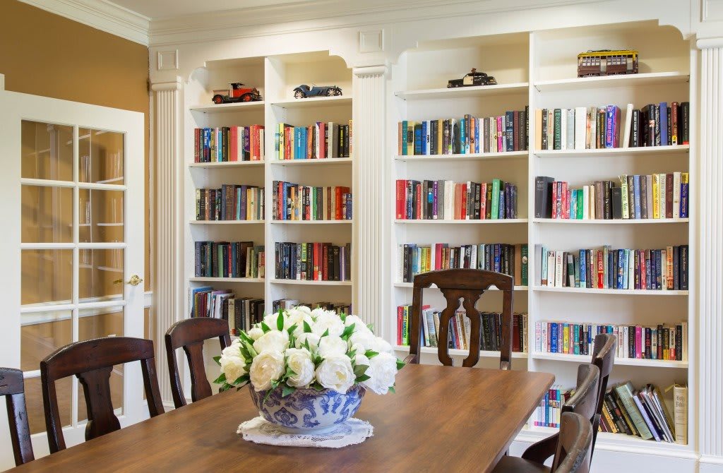 A cozy library room with white bookshelves filled with books and a wooden table adorned with a vase of flowers.