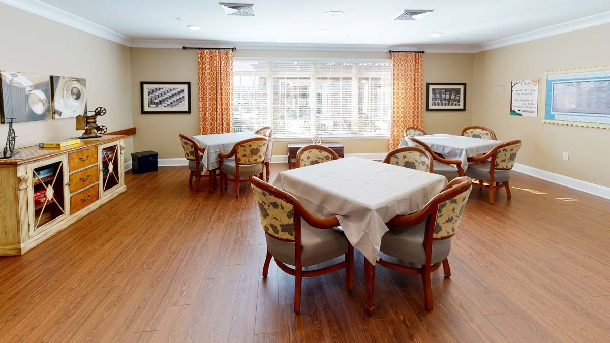 A bright dining room with several tables and chairs, featuring large windows and decorative curtains.