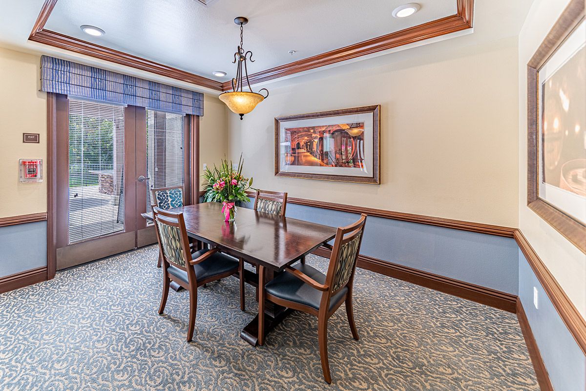 A cozy dining room with a wooden table and four chairs, featuring a chandelier, artwork on the wall, and a window letting in natural light.
