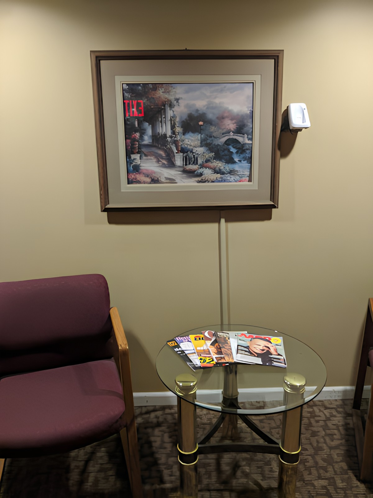 A waiting room with a chair, glass table holding magazines, and a painting on the wall.