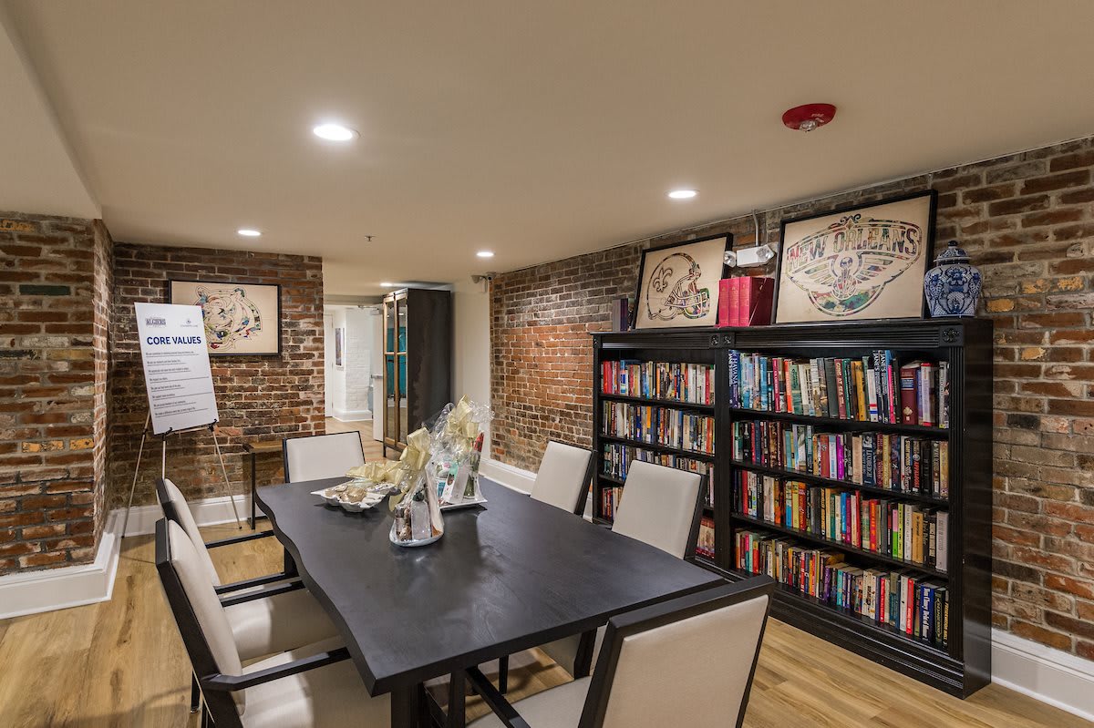 A cozy dining room with a large table, surrounded by chairs and decorated with bookshelves and artwork on exposed brick walls.