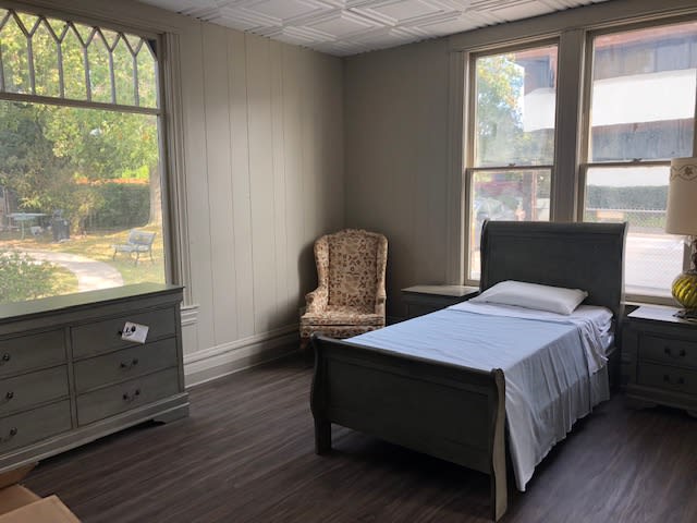 A minimalist bedroom with a simple bed, chair, dresser, and large windows.