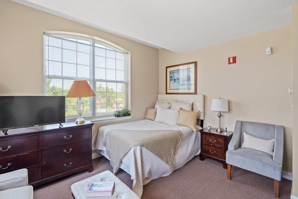 A cozy bedroom with a large window, featuring a bed, dresser, lamps, and seating area.