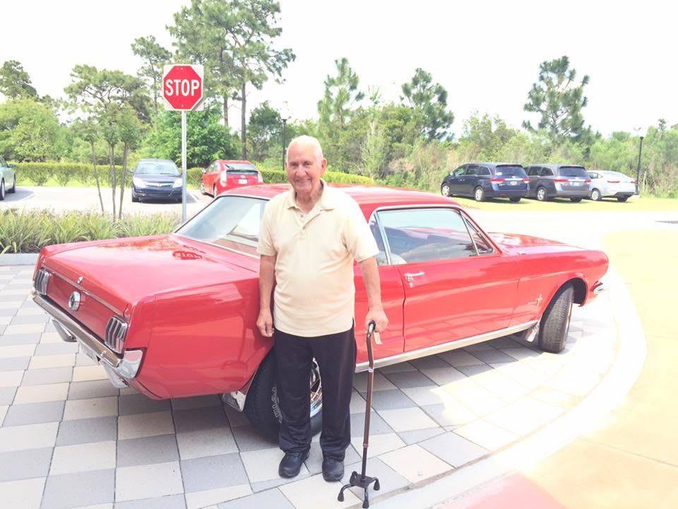 A man stands with a cane beside a classic red car in an outdoor parking area.