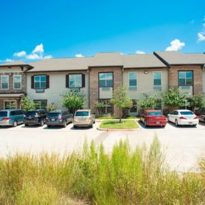A sunny day view of a residential apartment complex with a parking lot filled with cars.