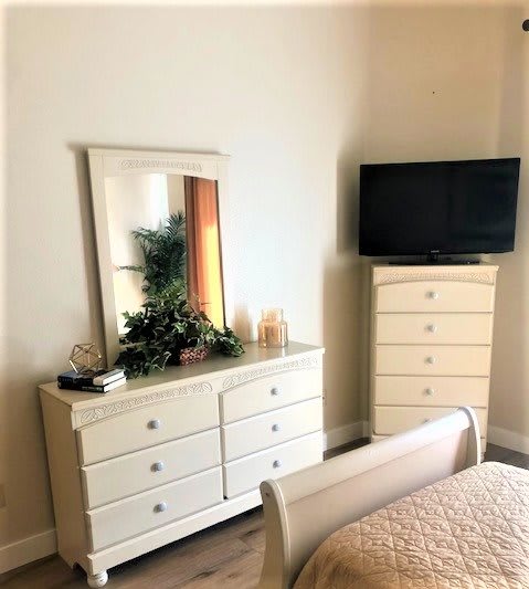 A cozy bedroom features a white dresser with a mirror and a TV on top of a tall chest of drawers.