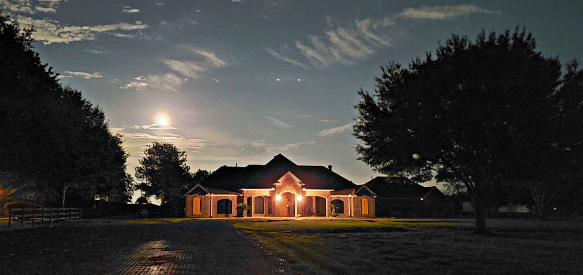 A house illuminated by outdoor lights under a night sky with clouds and the moon.