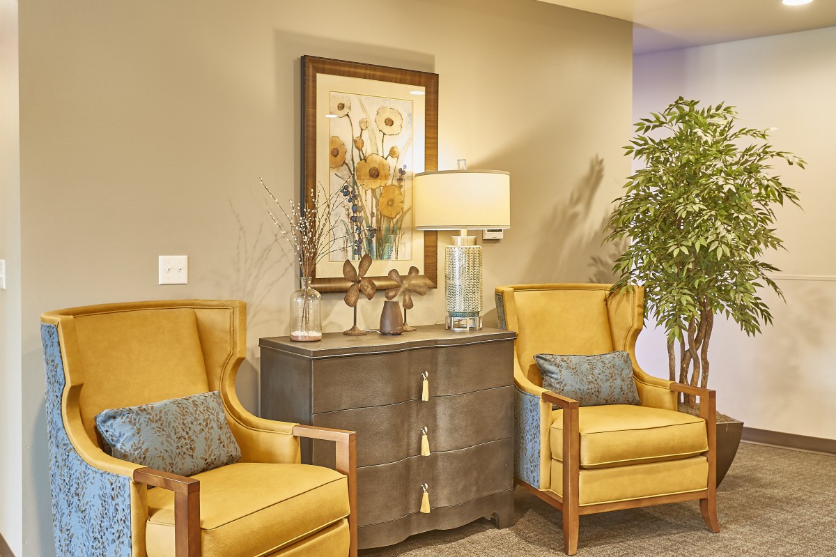 A cozy lounge area featuring two yellow chairs, a central cabinet with a lamp, and decorative elements such as a painting and indoor plant.