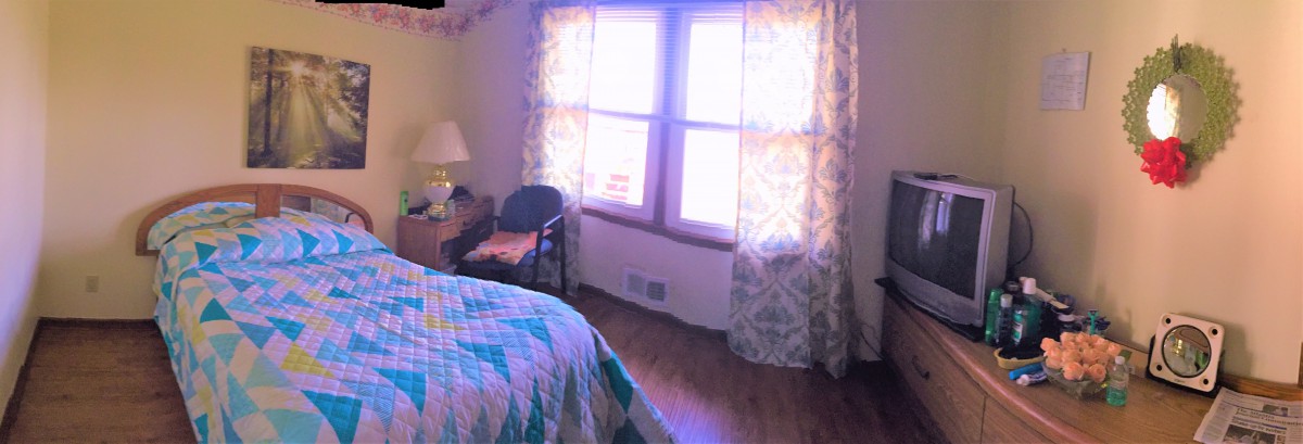 A cozy bedroom with a quilt-covered bed, a chair, and patterned curtains framing a sunlit window.