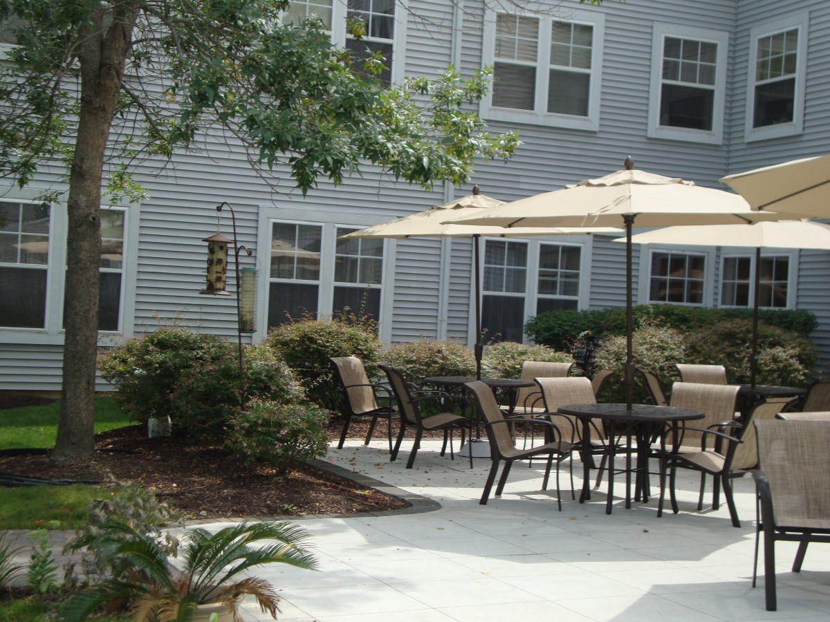 A cozy outdoor patio area with tables, chairs, and umbrellas, surrounded by bushes and adjacent to a building.