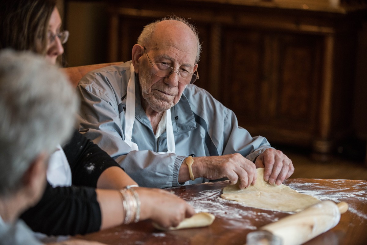 An elderly man wearing an apron, working with dough in a kitchen setting.