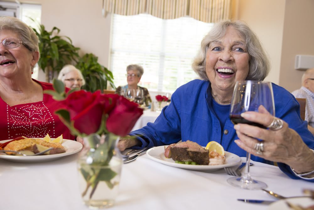 Elderly women enjoying a meal together at a dining table, with one woman smiling and holding a wine glass.