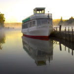 A boat docked on a calm, misty morning with its reflection visible in the water.