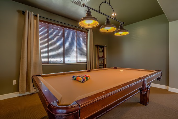A cozy game room featuring a pool table with overhead lighting and window blinds.