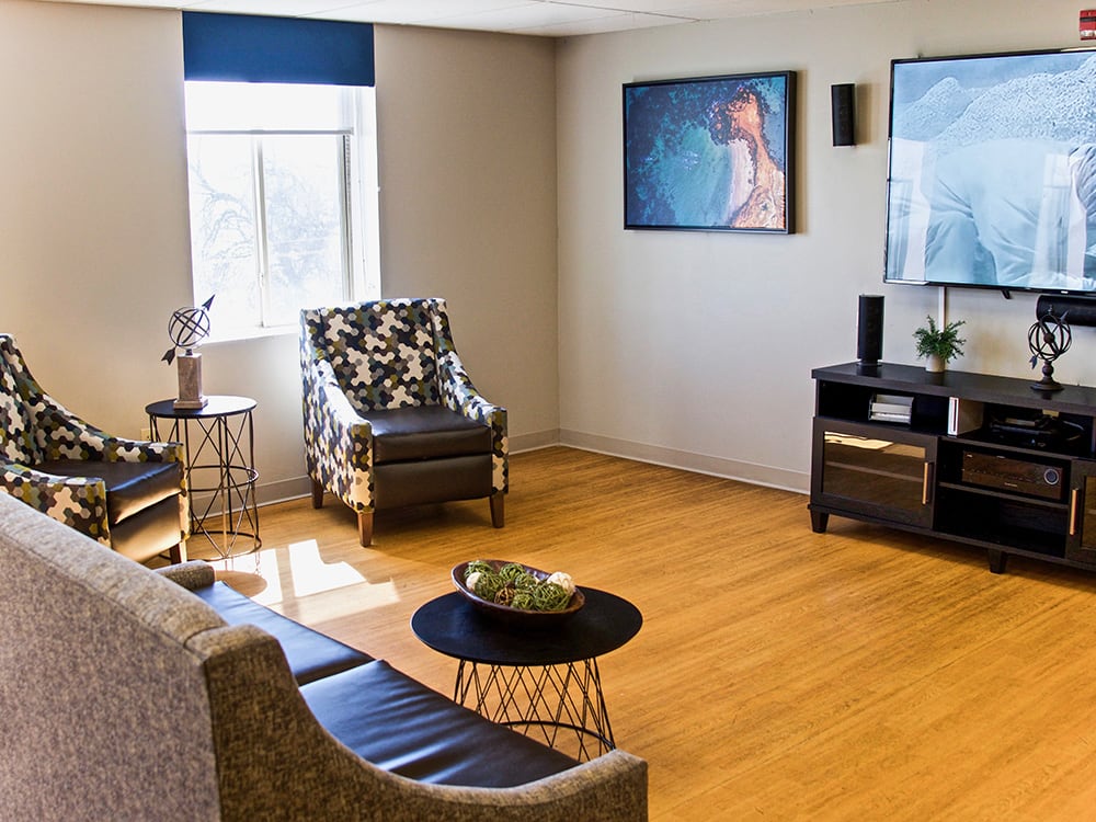 A modern living room with wooden flooring, featuring a sofa, armchair, and a TV on the wall.