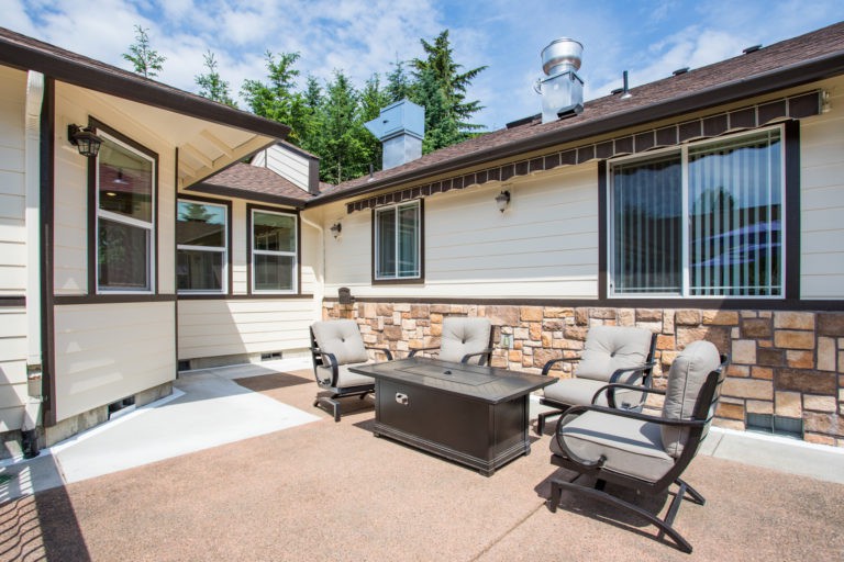 A cozy patio with outdoor seating and a table, set against the backdrop of a modern house exterior on a sunny day.