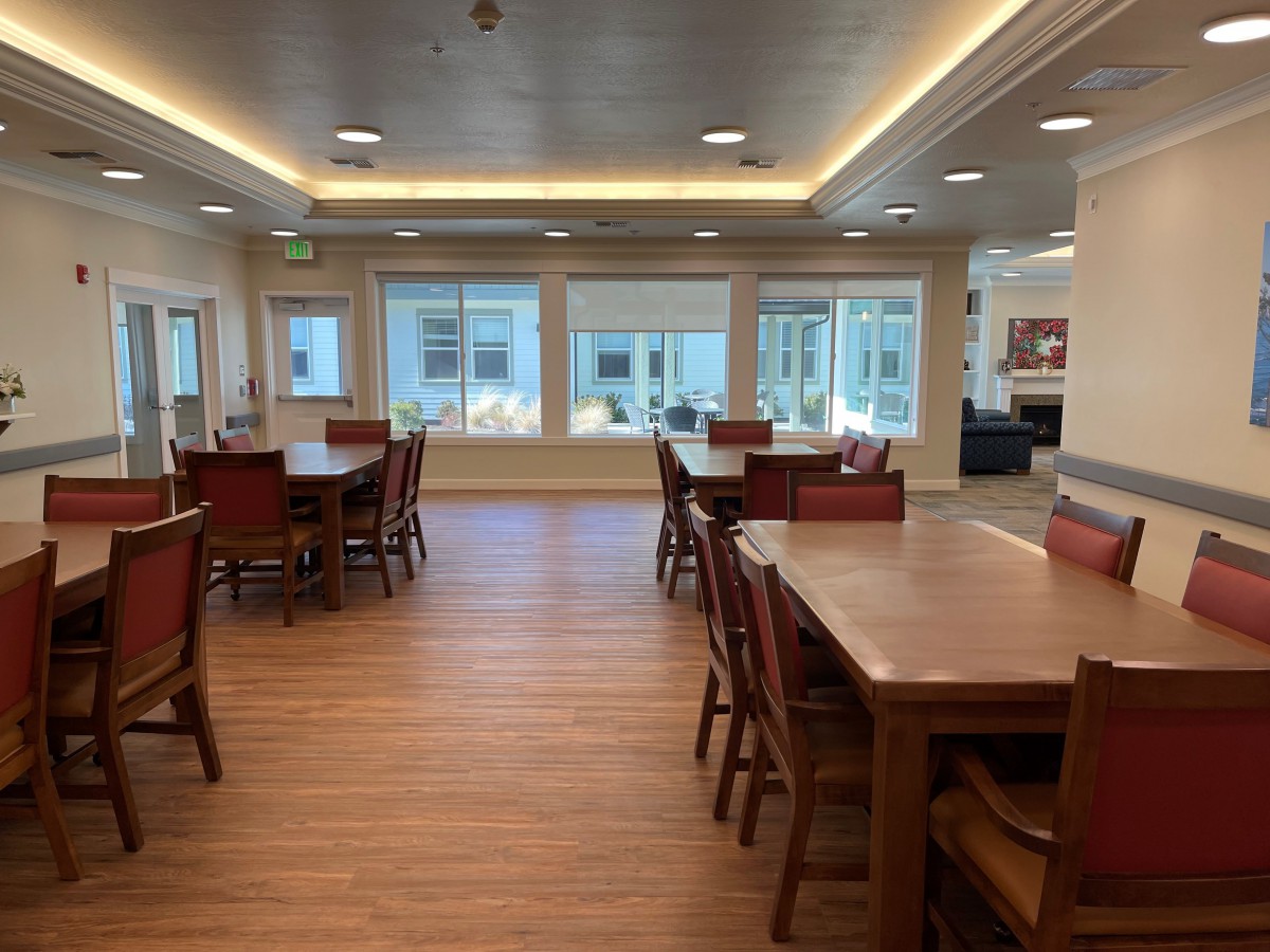 A spacious dining area with wooden tables and chairs, illuminated by ceiling lights and natural light from large windows.