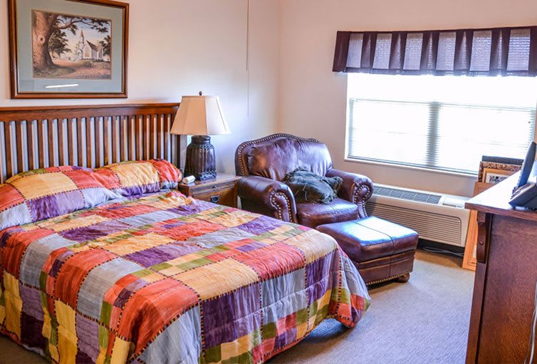 A cozy bedroom with a colorful quilt on the bed, an armchair by the window, and a lamp on the bedside table.
