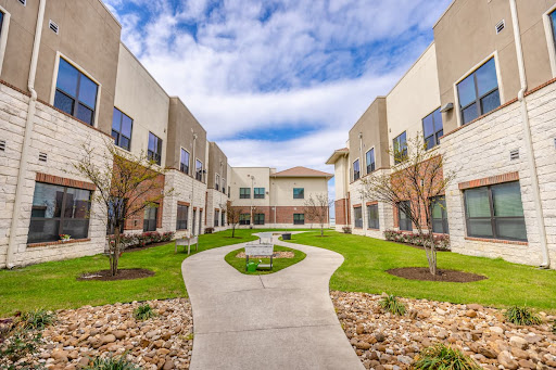 A serene courtyard surrounded by modern buildings, featuring a winding pathway and lush green grass under a partly cloudy sky.