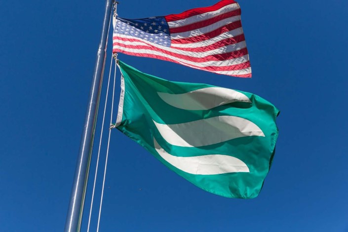 An American flag flying next to a green flag with two white diagonal stripes against a clear blue sky.