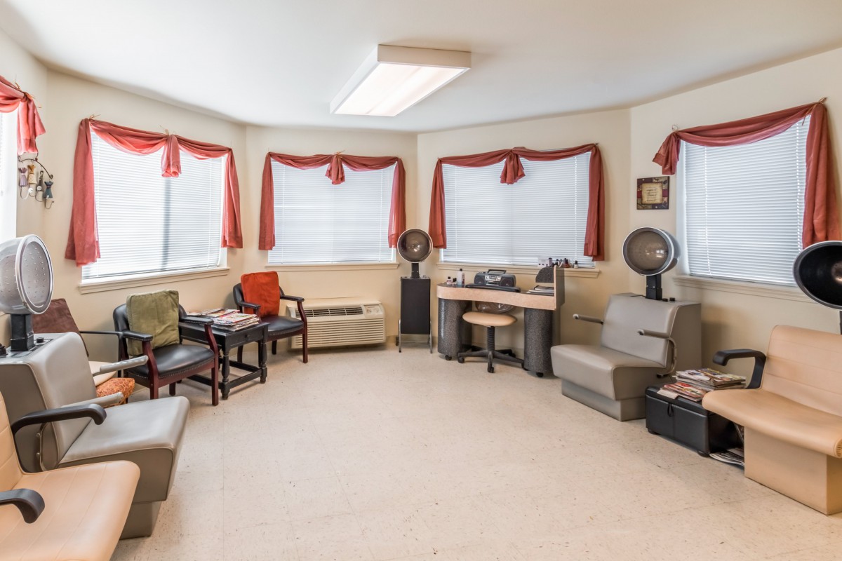A cozy salon waiting area with chairs, hair dryers, and bright windows adorned with red curtains.