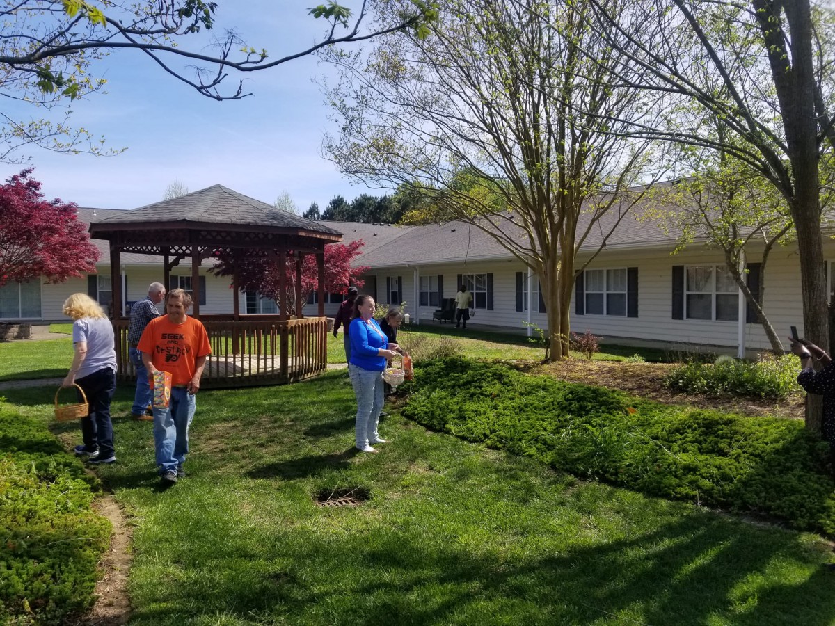 A group of people walking in a garden area with a gazebo, trees, and a building in the background on a sunny day.