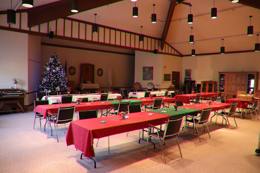 A spacious event room set up for a holiday gathering, featuring red and green tablecloths and a decorated Christmas tree.