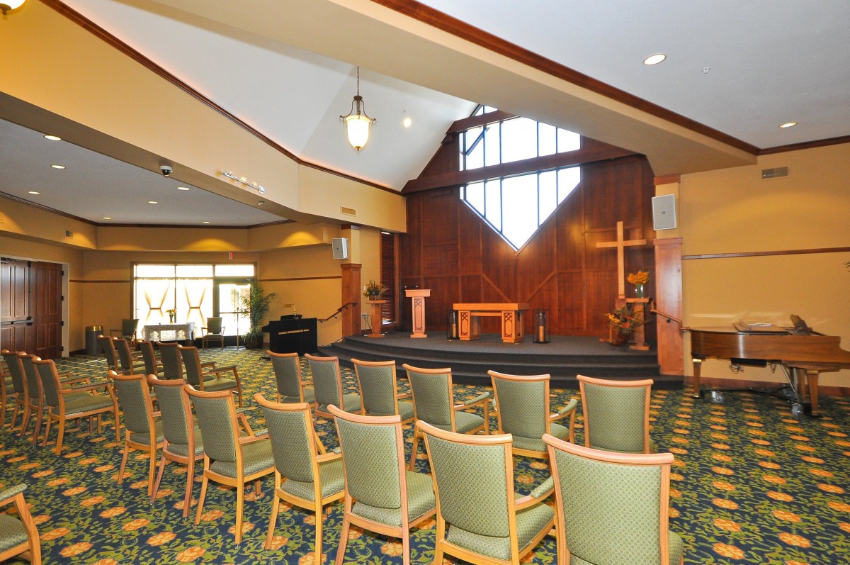 A chapel interior with rows of chairs, a podium on stage, and a large stained glass window.