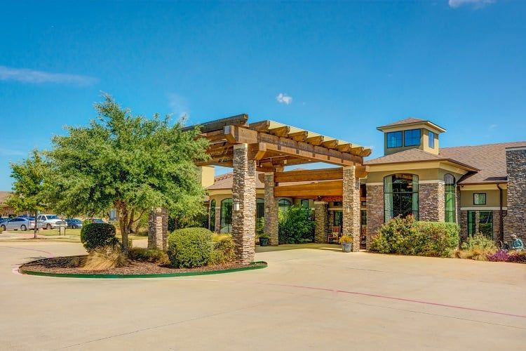 A sunny day view of a building entrance with a pergola and landscaping.