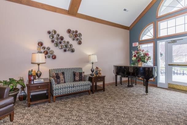 A cozy living room with a sofa, coffee tables, lamps, and a piano, featuring elegant wall art and carpeted flooring.