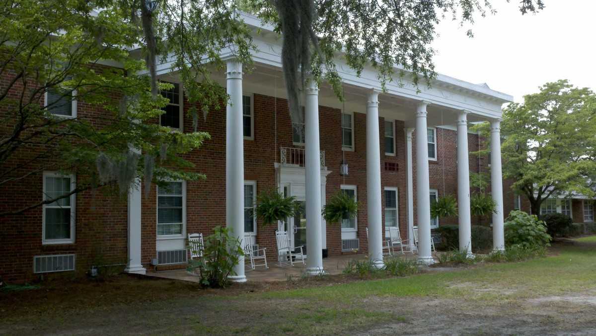 A historic brick building with large white columns on the porch, surrounded by trees.