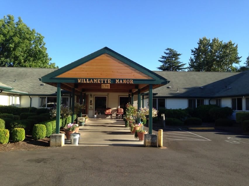 A welcoming entrance to a building with a sign reading 'Willamette Manor', surrounded by trees and a parking lot.