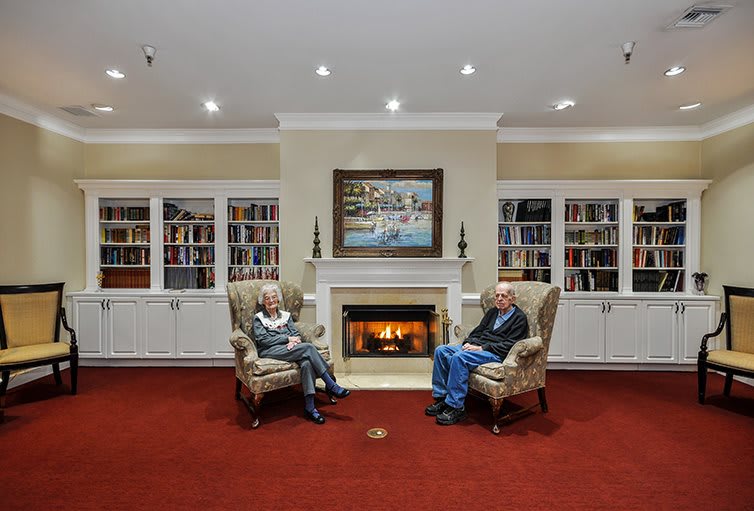 Two people sitting in armchairs by a fireplace in a library setting.