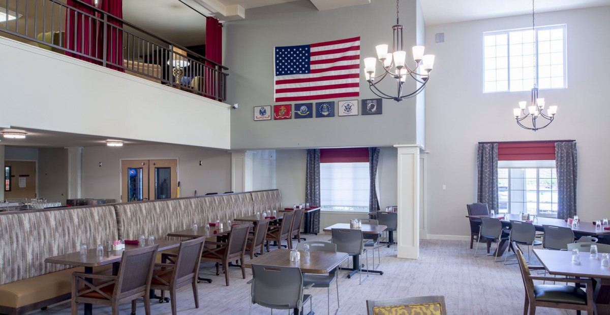 A spacious dining area featuring an American flag and military emblems on the wall, with a modern chandelier and neatly arranged tables and chairs.