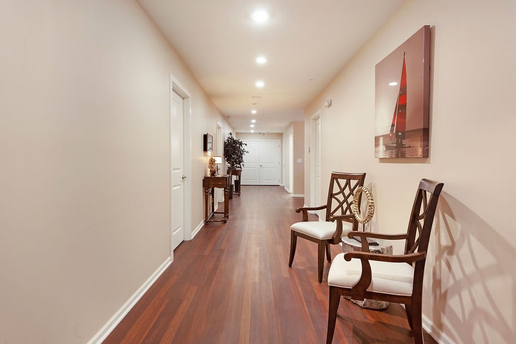 A well-lit hallway with wooden flooring, featuring decorative chairs and artwork on the walls.