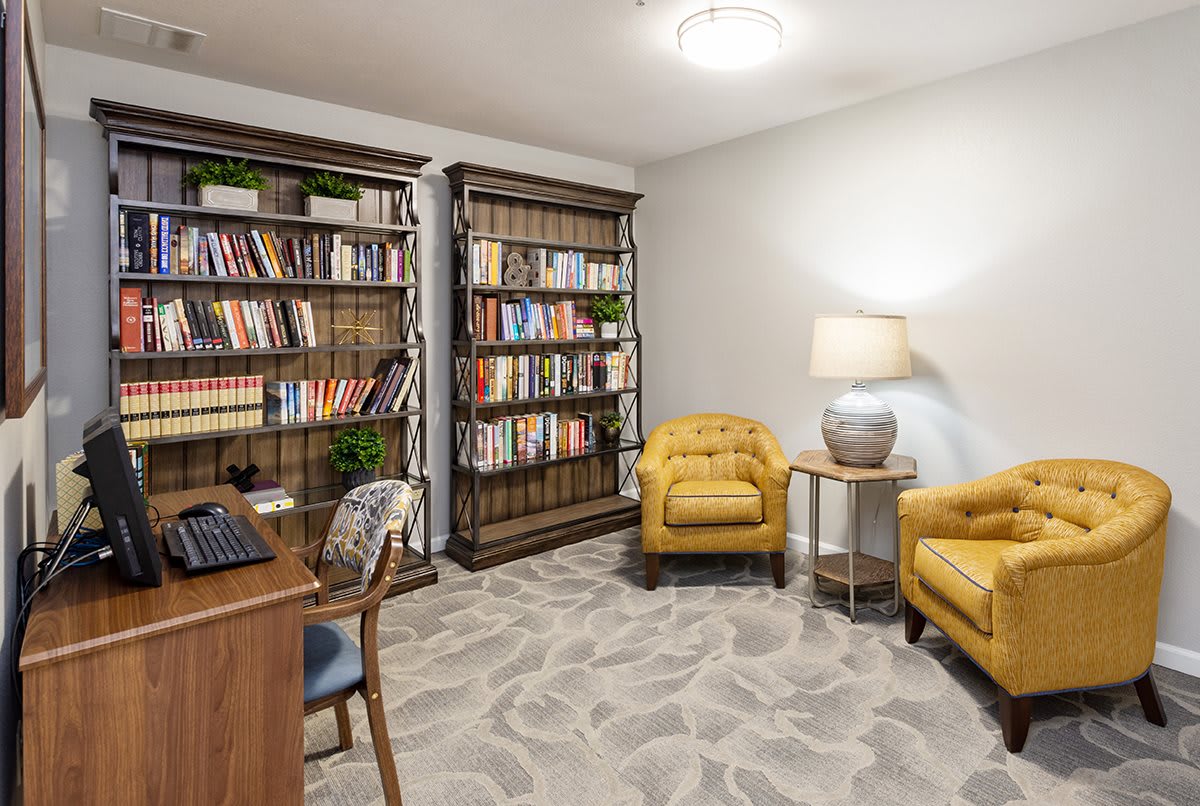 A cozy home office featuring a desk, bookshelves filled with books, and a seating area with two yellow armchairs and a lamp.