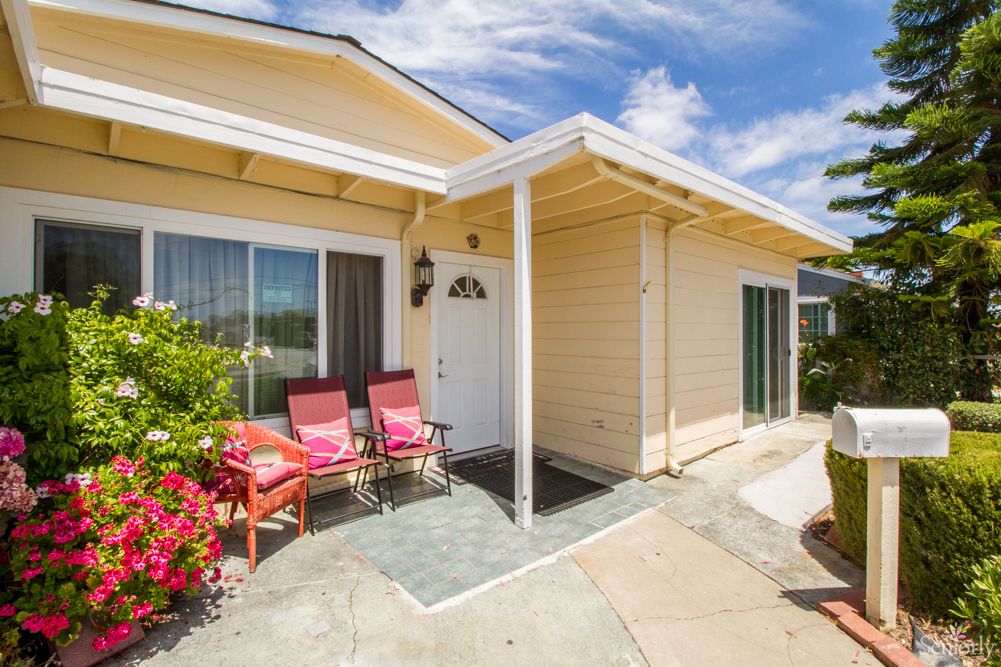A sunny view of a house exterior with a cozy front porch featuring two red chairs and surrounding greenery.