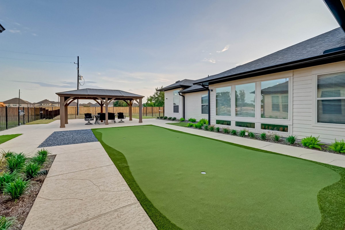A modern backyard with a putting green, patio, and pergola adjacent to a house.