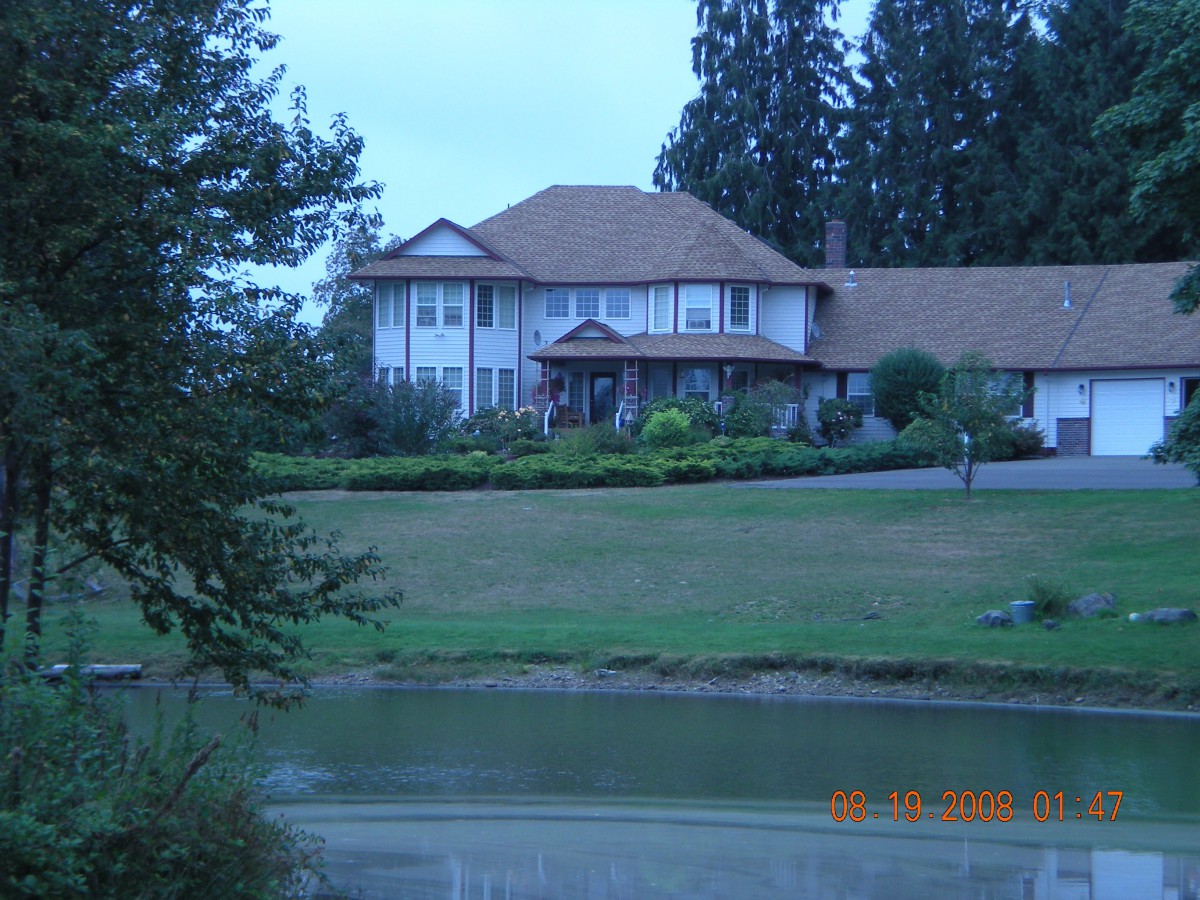 A large house with a sloped roof, surrounded by trees and greenery, overlooks a pond.