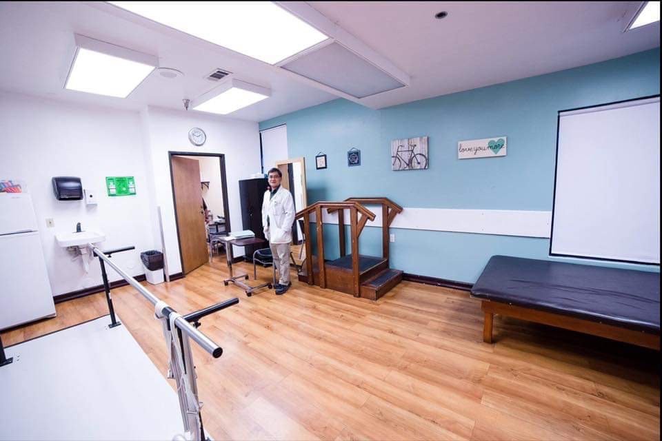 A physical therapy room equipped with exercise equipment, including parallel bars and a treadmill, with a healthcare professional present.