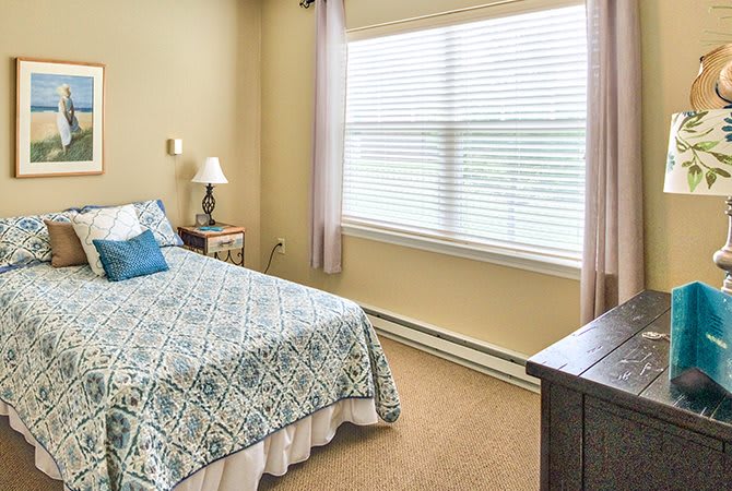 A cozy bedroom featuring a neatly made bed with patterned bedding, a window with blinds, and a bedside lamp.