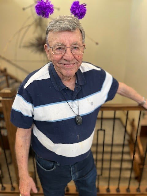 An elderly man wearing a striped shirt and glasses stands by a staircase, smiling.